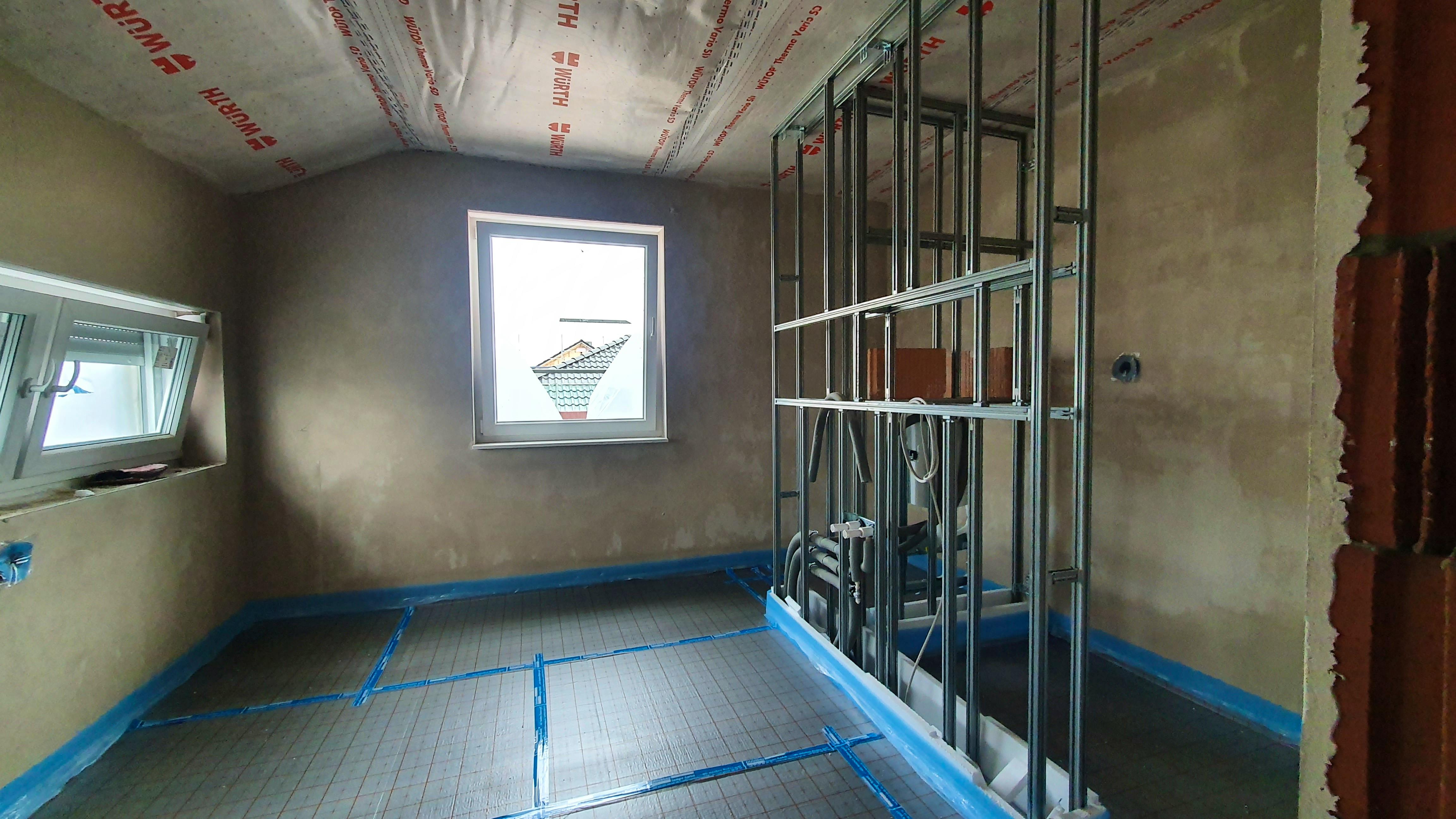 Zimmer im Rohbau-Innenausbau mit verputzten Wänden, Fußbodenheizung, Metallständerwerk für eine Trockenbauwand und einem quer angeordneten Fenster über der geplanten Badewannen-Position