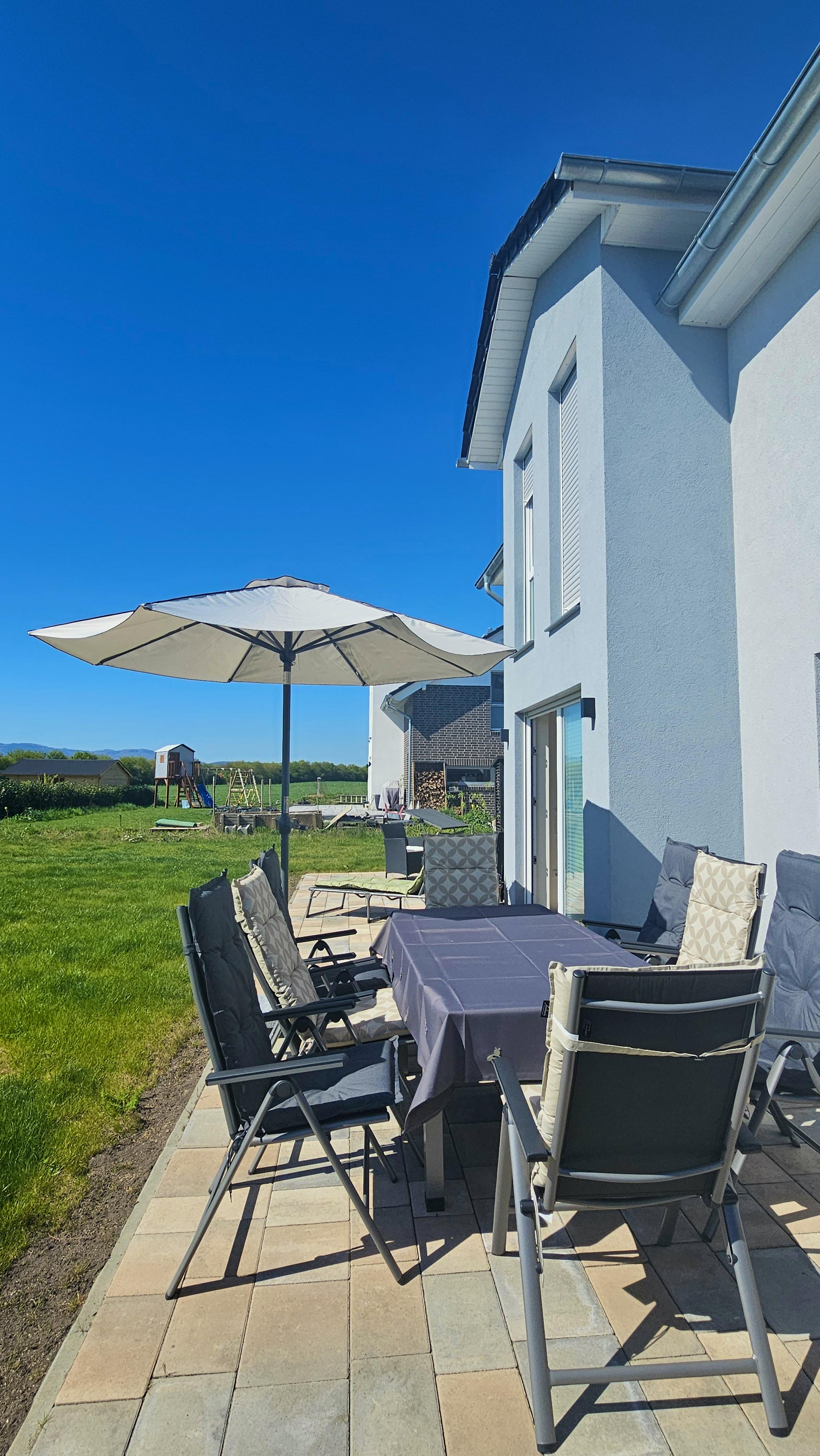 Gepflasterte Terrasse mit Sonnenschirm, Gartentisch und Stühlen an der Hausseite mit Standerker; im Hintergrund freie Feldlage mit Blick auf den Harz