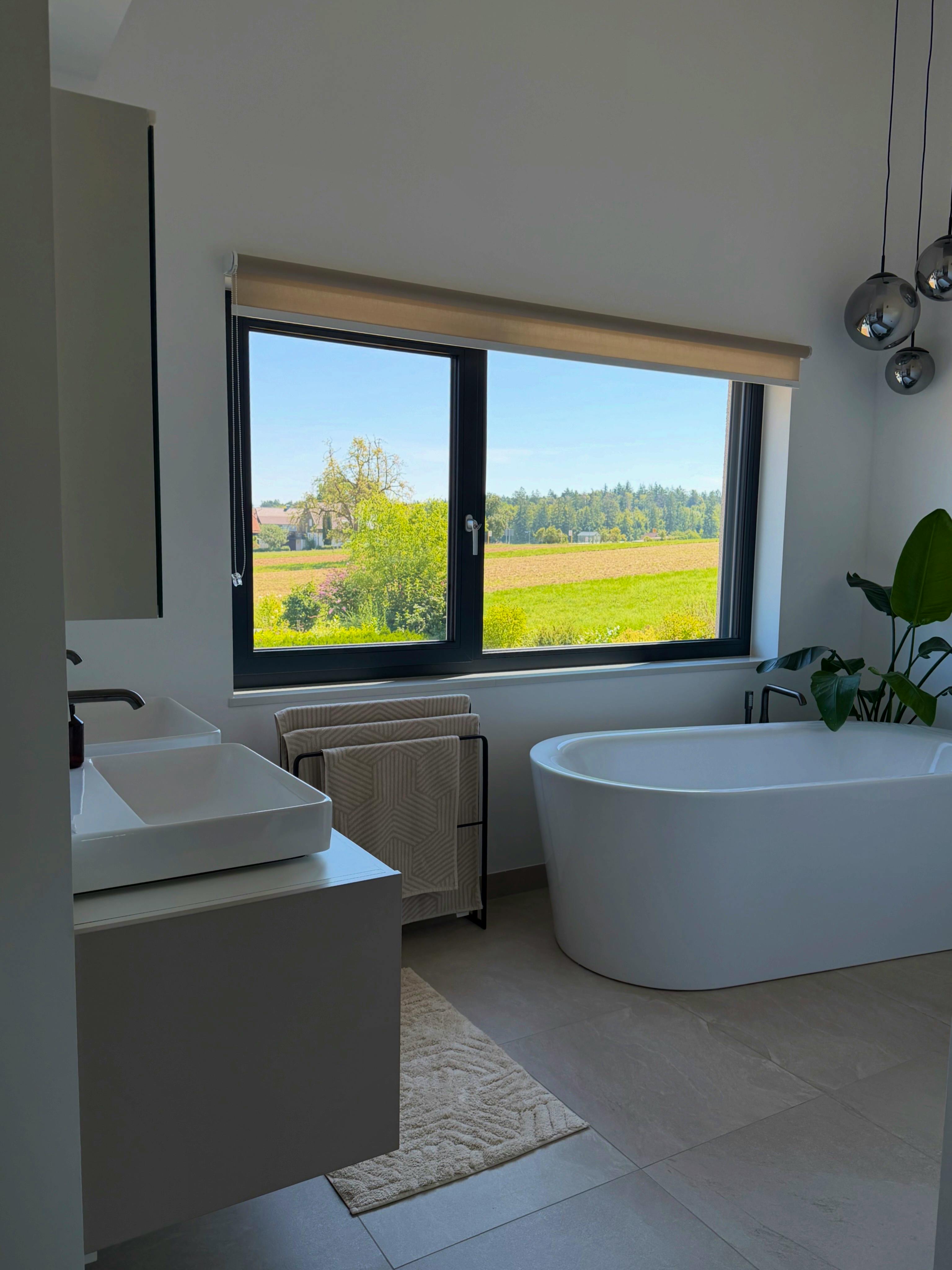 Hauptbadezimmer mit Doppelwaschtisch, freistehender Badewanne, großem Fenster mit Blick auf Felder und Rauchglas-Pendelleuchten