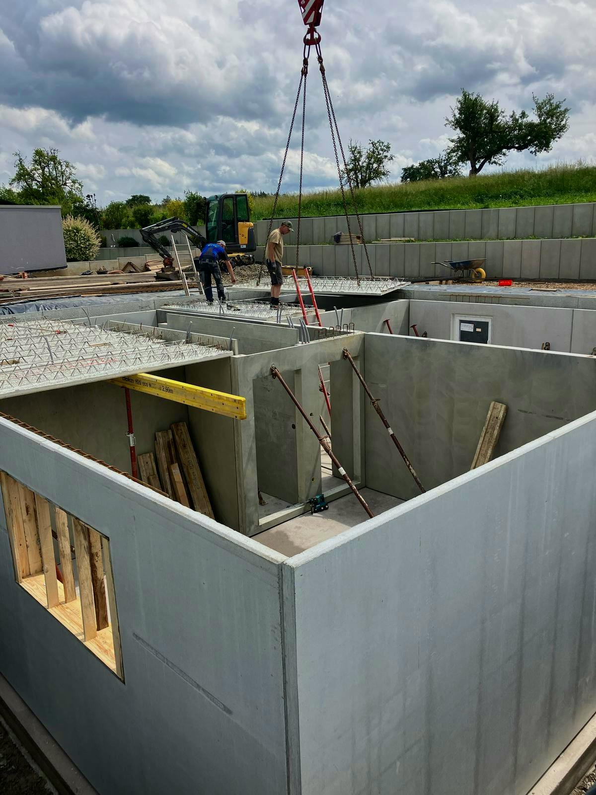 Fertigkeller im Aufbau: Betonwände stehen, ein Wandelement wird per Kran eingesetzt, zwei Arbeiter stehen auf der Kellerdecke, im Hintergrund grüne Felder