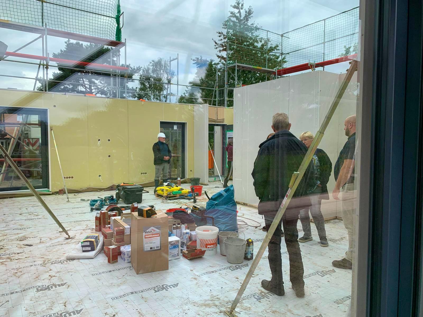 Blick durch Fensterfront in den noch offenen Rohbau eines Kampa-Fertighauses während der Aufstellung mit Stelltrupp und Materialvorbereitung