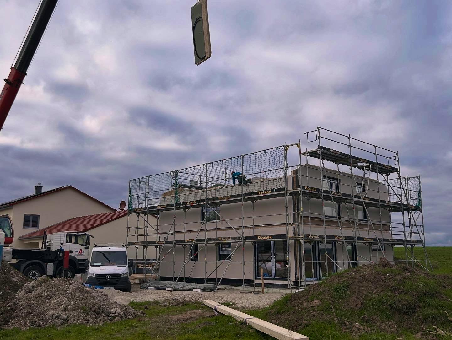 Haustelltag des Hanse-Haus-Fertighauses – Kran hebt Dachelement auf das eingerüstete Obergeschoss, Montagefahrzeuge auf dem Grundstück.