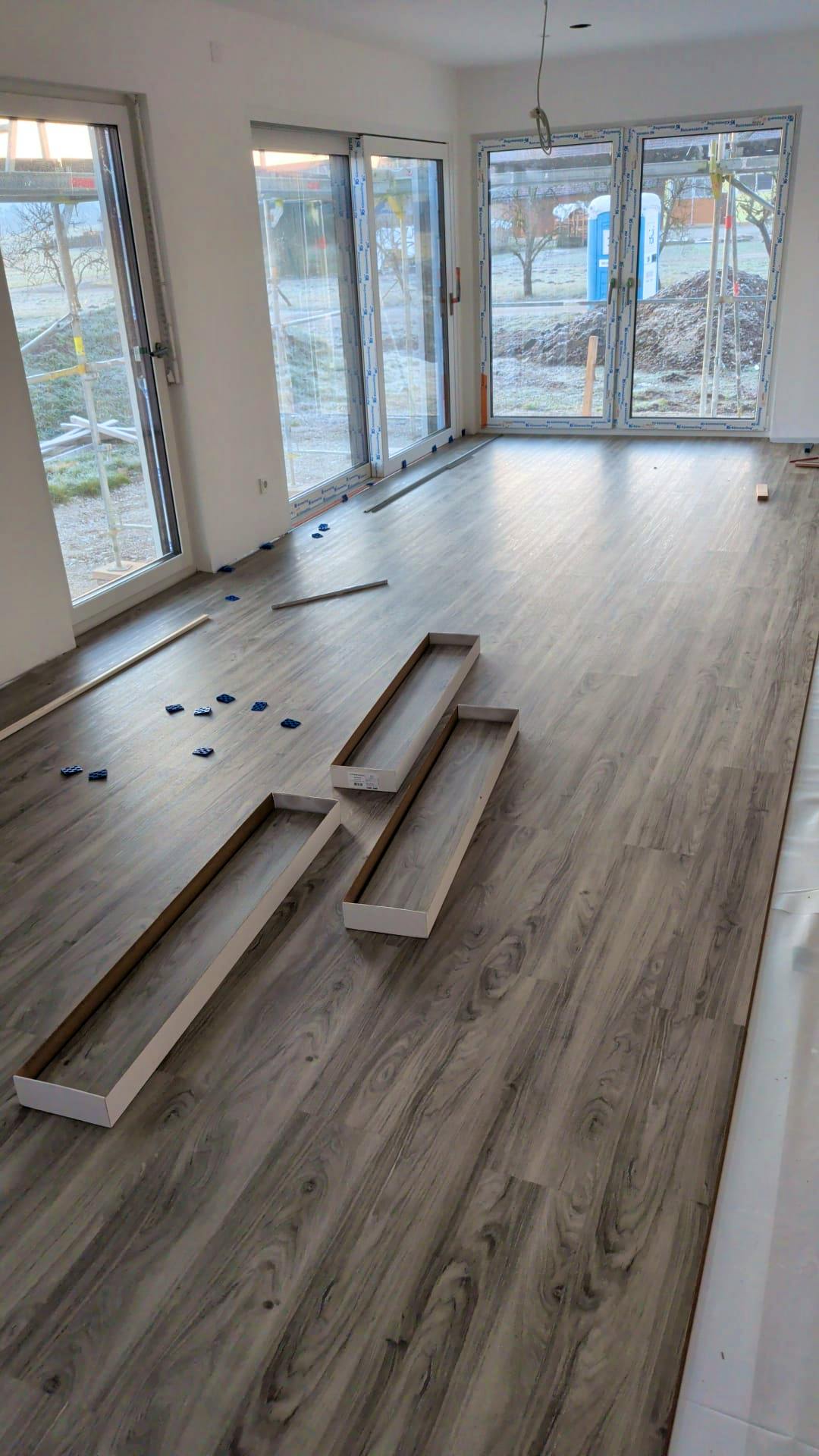 Eigenleistung im Hanse-Haus-Fertighaus – Vinylboden wird im Wohnbereich verlegt, Fußleisten liegen bereit, bodentiefe Fenster im Hintergrund.