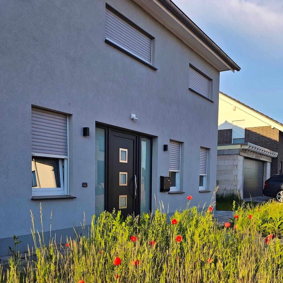 Zweigeschossiges Massivhaus mit hellgrauem Putz, schwarzer Haustür mit goldenen Details, geschlossenen Rollläden und einer Wildblumenwiese im Vordergrund