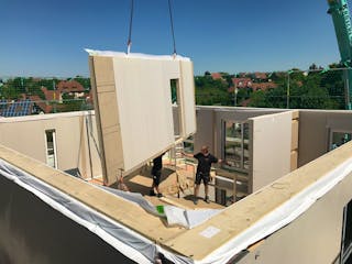 Aufstellung eines Fertighauses: Vorgefertigte Wandelemente werden auf der Bodenplatte montiert, im Hintergrund bereits bezogene Nachbarhäuser