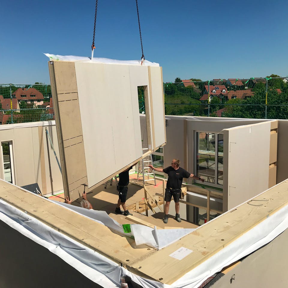 Aufstellung eines Fertighauses: Vorgefertigte Wandelemente werden auf der Bodenplatte montiert, im Hintergrund bereits bezogene Nachbarhäuser