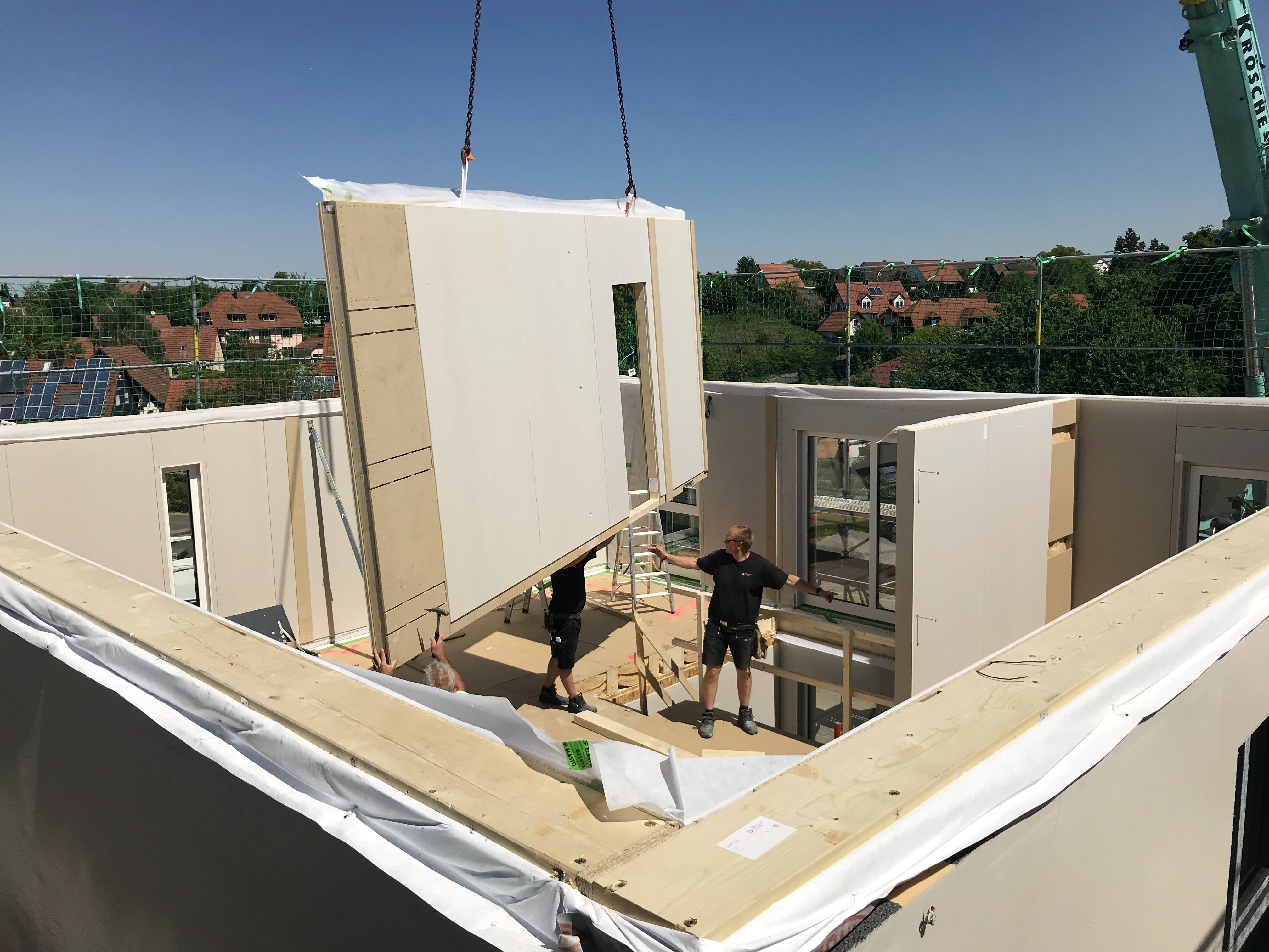 Aufstellung eines Fertighauses: Vorgefertigte Wandelemente werden auf der Bodenplatte montiert, im Hintergrund bereits bezogene Nachbarhäuser