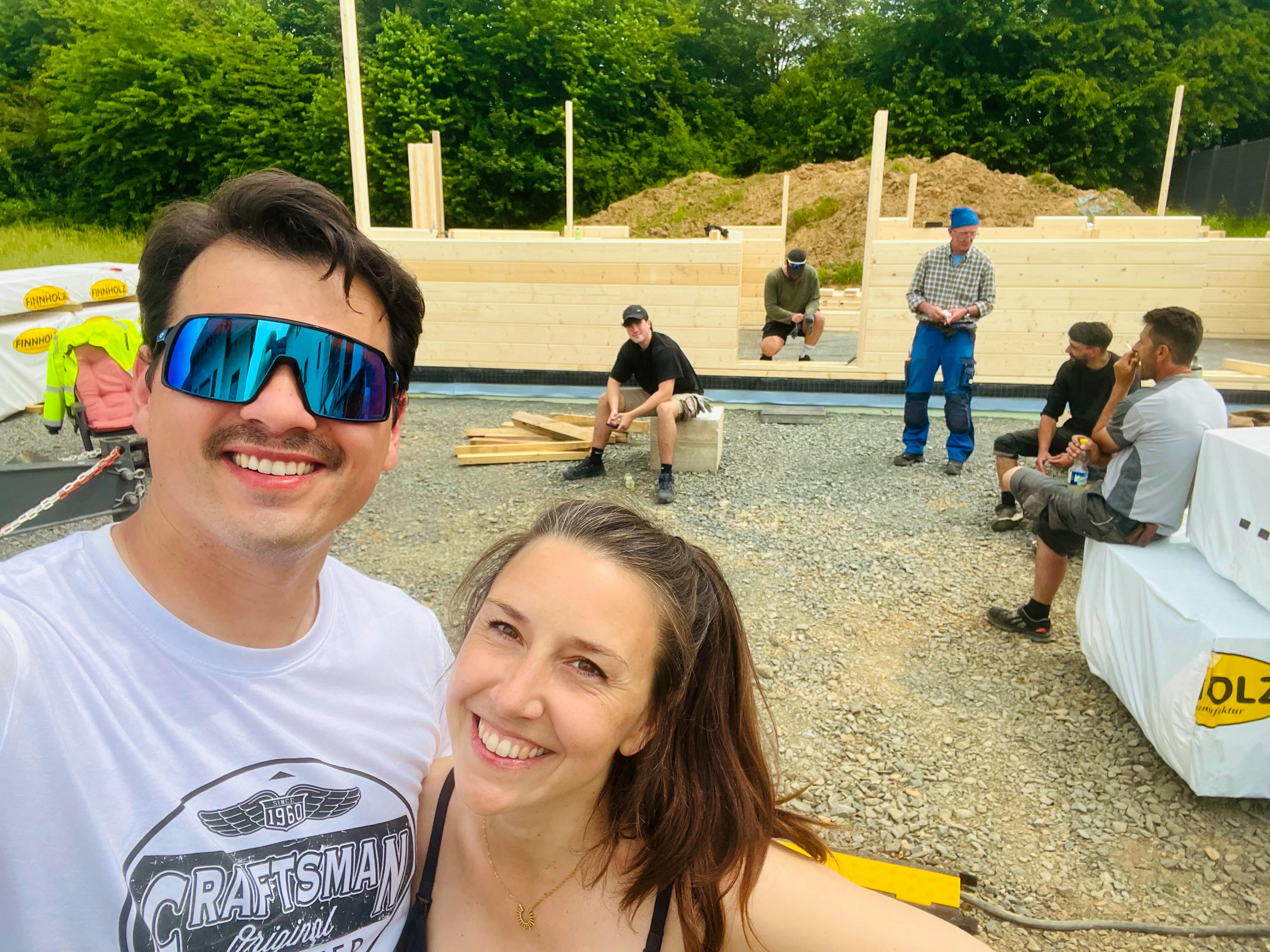 Tim und Annemarie Ellrich lächeln auf ihrer Blockhaus-Baustelle, im Hintergrund die ersten Blockbohlenlagen und Helfer bei der Pause