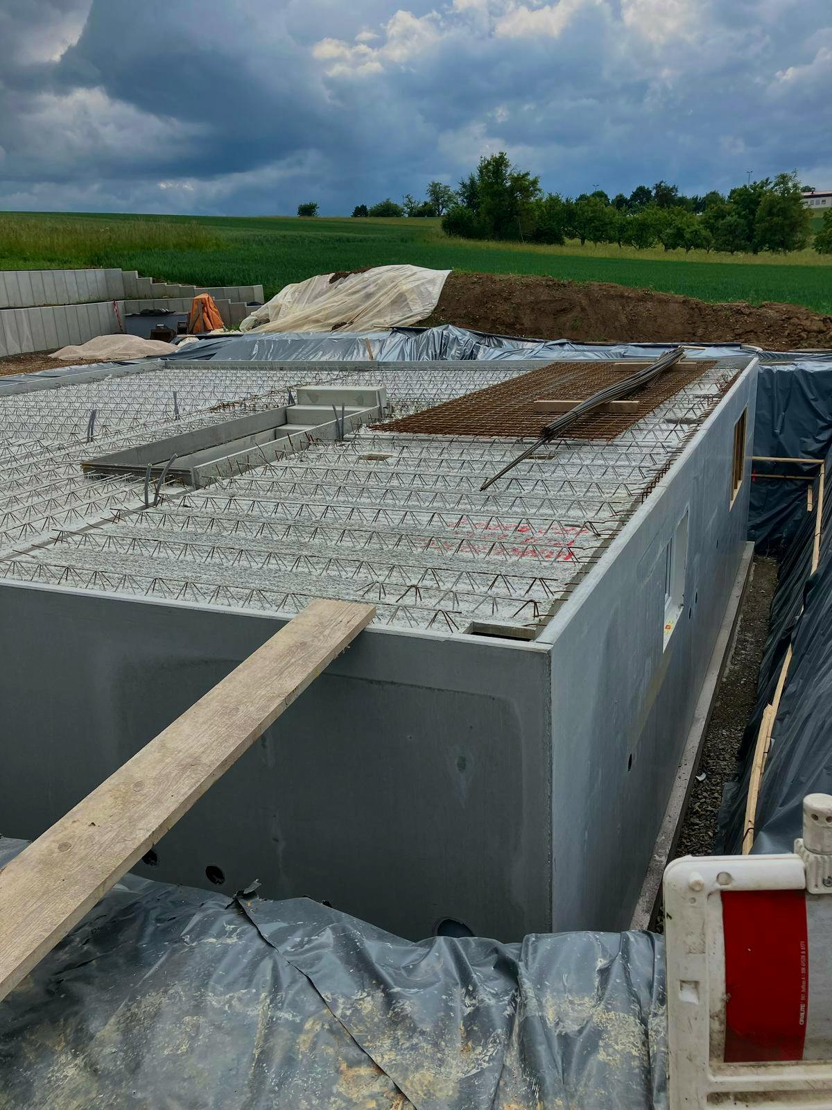 Fertigkeller im Rohbau: Betonfertigwände mit aufgelegten Filigrandeckenelementen und Bewehrung, bereit zum Betonieren der Kellerdecke. Außen ist die schwarze Abdichtungsbahn sichtbar. Baustelle auf einem ländlichen Grundstück.