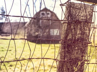 Ein Bauernhaus steht auf weitem Grundstück hinter einem Drahtzaun