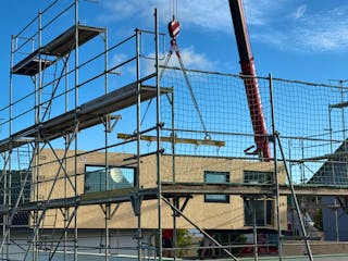 Baustelle eines Fertighauses in Holzrahmenbauweise: Ein Kran hebt vorgefertigte Wandelemente mit sichtbaren OSB-Platten auf das Obergeschoss, das Erdgeschoss ist bereits verputzt und verglast.