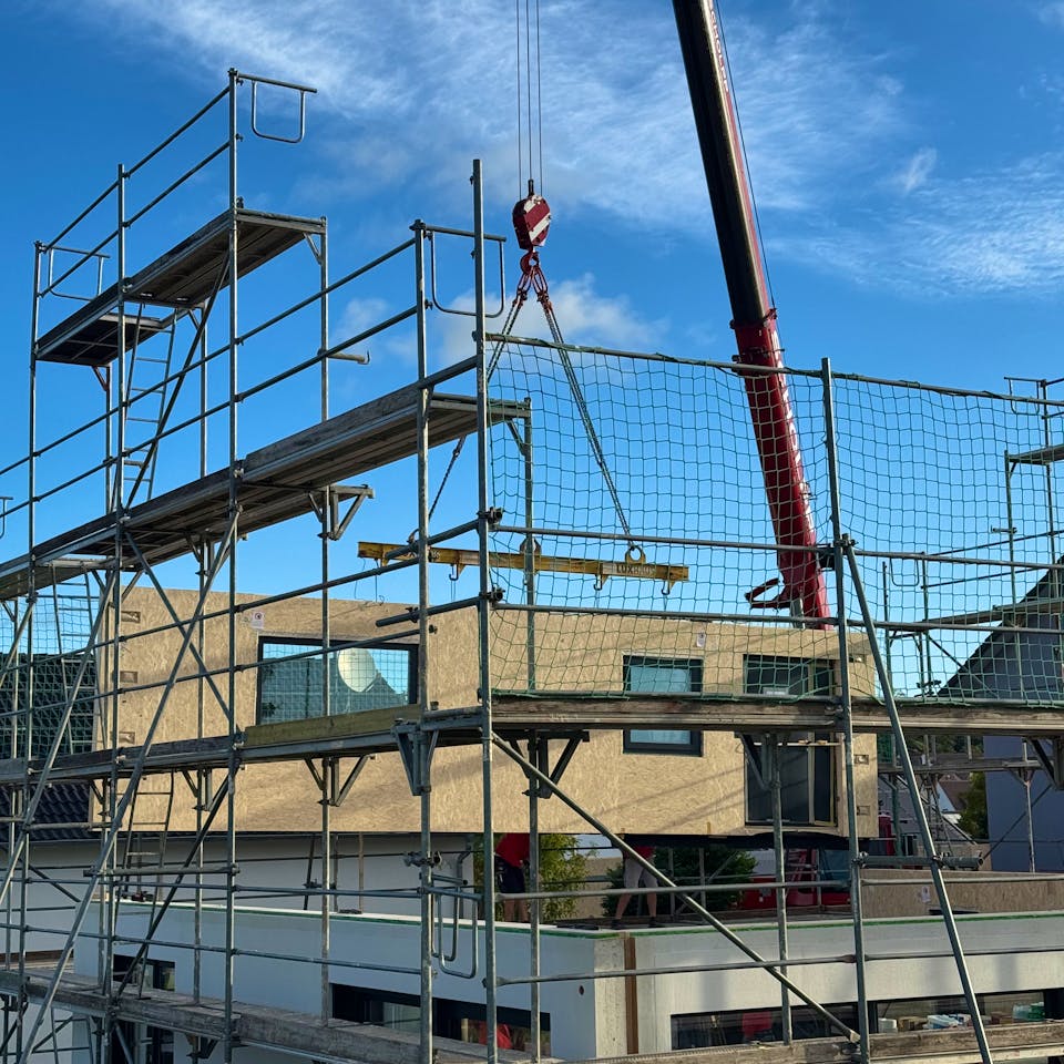 Baustelle eines Fertighauses in Holzrahmenbauweise: Ein Kran hebt vorgefertigte Wandelemente mit sichtbaren OSB-Platten auf das Obergeschoss, das Erdgeschoss ist bereits verputzt und verglast.