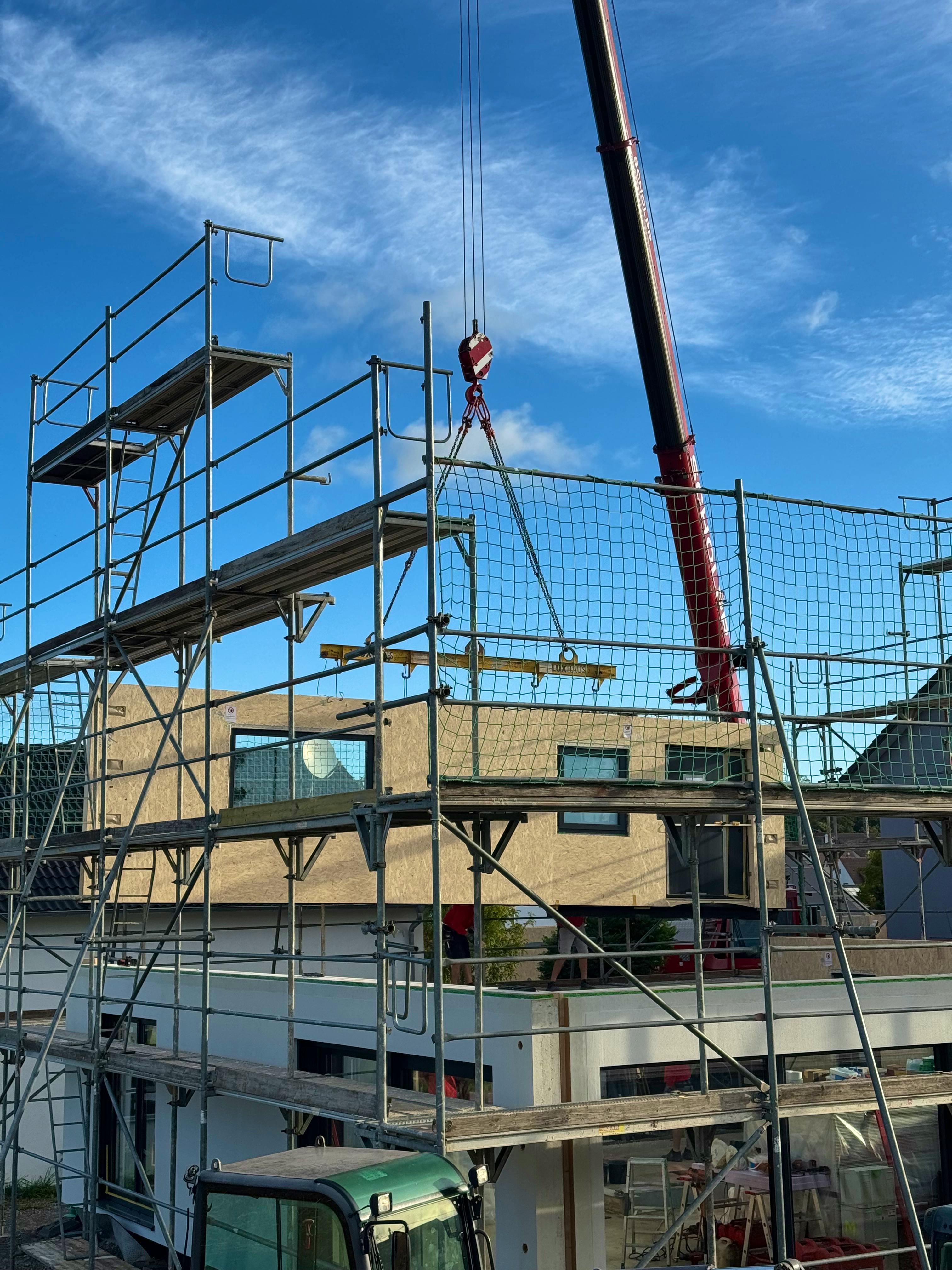 Baustelle eines Fertighauses in Holzrahmenbauweise: Ein Kran hebt vorgefertigte Wandelemente mit sichtbaren OSB-Platten auf das Obergeschoss, das Erdgeschoss ist bereits verputzt und verglast.
