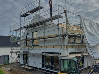 Zweistöckiges Fertighaus im Rohbau mit Baugerüst und Kranmontage, davor ein Minibagger auf der Baustelle – Bauprojekt einer jungen Familie im Raum Stuttgart.