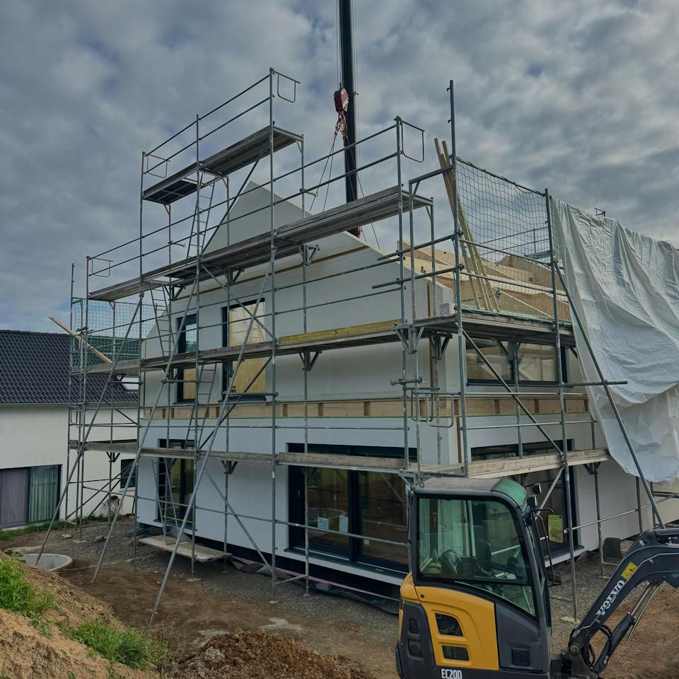 Zweistöckiges Fertighaus im Rohbau mit Baugerüst und Kranmontage, davor ein Minibagger auf der Baustelle – Bauprojekt einer jungen Familie im Raum Stuttgart.