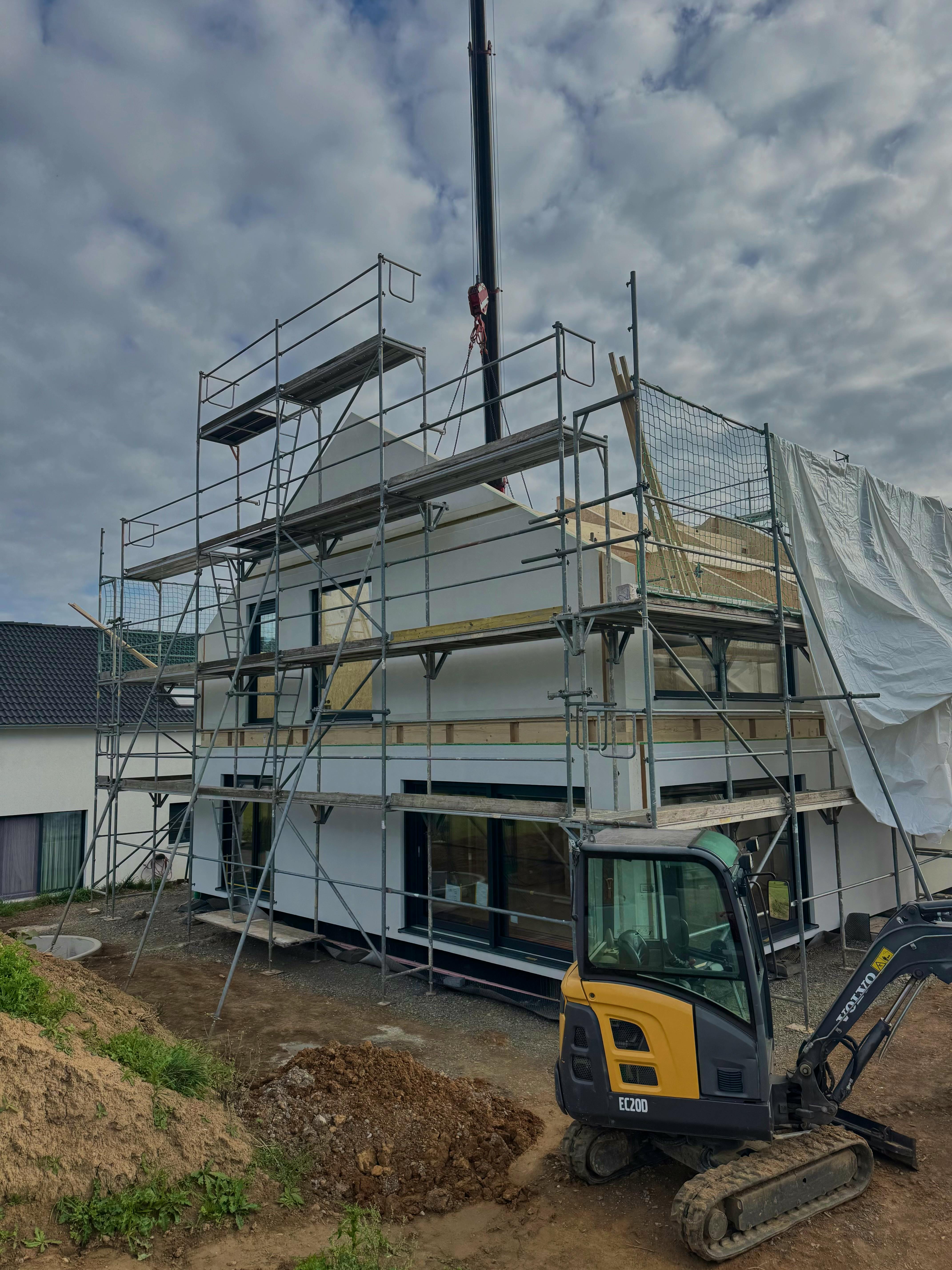 Zweistöckiges Fertighaus im Rohbau mit Baugerüst und Kranmontage, davor ein Minibagger auf der Baustelle – Bauprojekt einer jungen Familie im Raum Stuttgart.