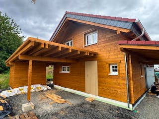 Fertiggestelltes Holzblockhaus bei Melsungen in Hessen mit Carport, Veranda, PV-Anlage auf dem Dach und Nachbarhaus im Hintergrund