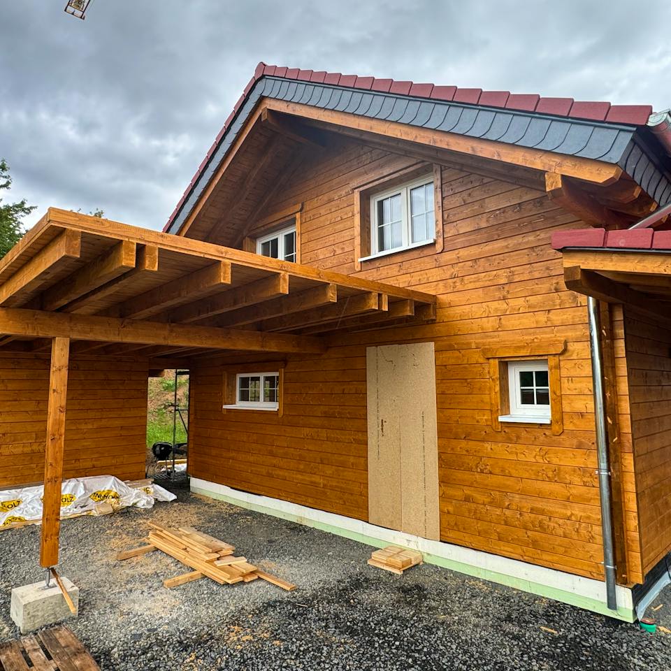 Fertiggestelltes Holzblockhaus bei Melsungen in Hessen mit Carport, Veranda, PV-Anlage auf dem Dach und Nachbarhaus im Hintergrund