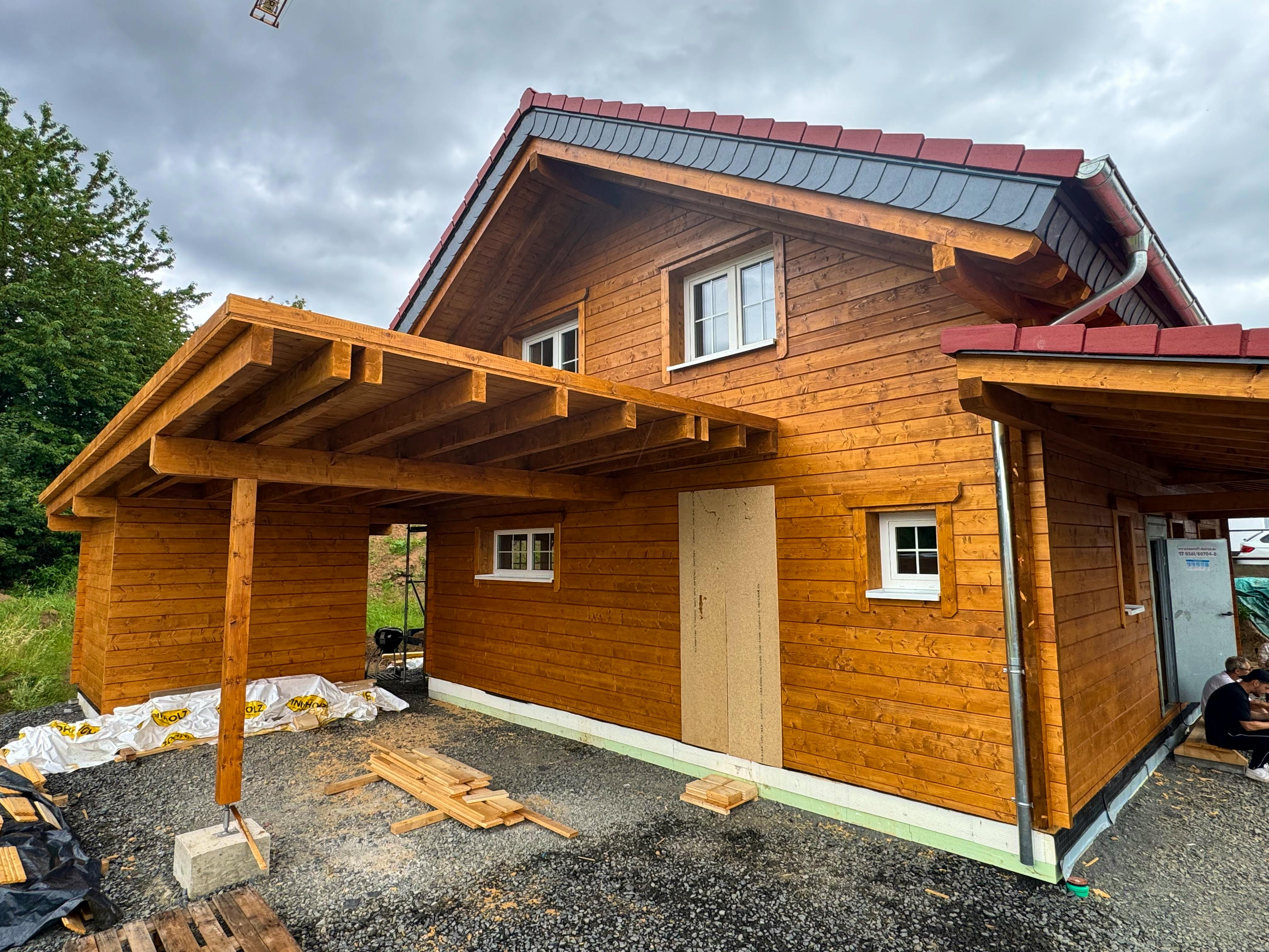 Fertiggestelltes Holzblockhaus bei Melsungen in Hessen mit Carport, Veranda, PV-Anlage auf dem Dach und Nachbarhaus im Hintergrund