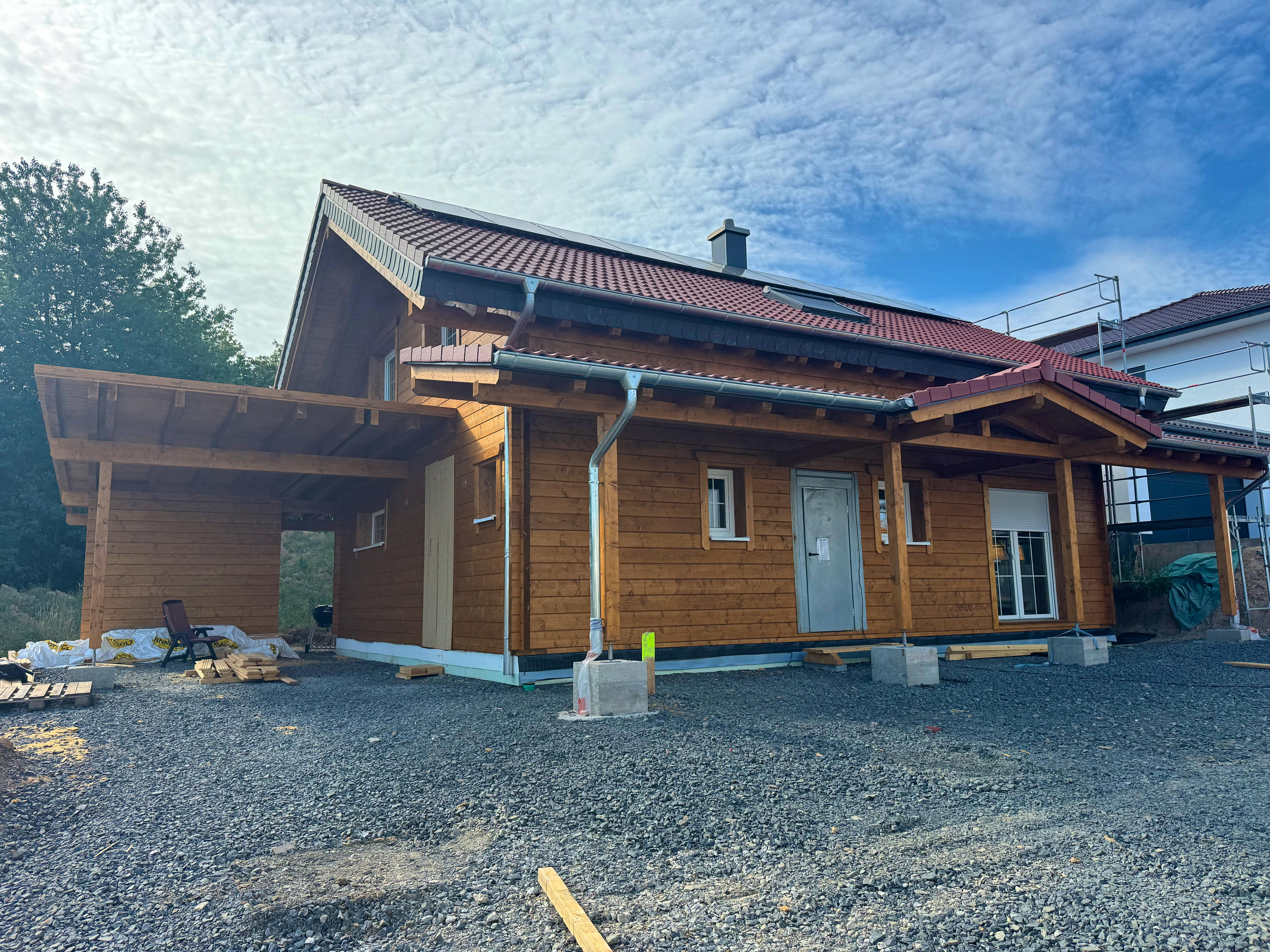 Gesamtansicht des fertigen Holzblockhauses bei Melsungen mit Carport, Veranda, PV-Anlage auf dem Dach und Nachbarhaus im Hintergrund