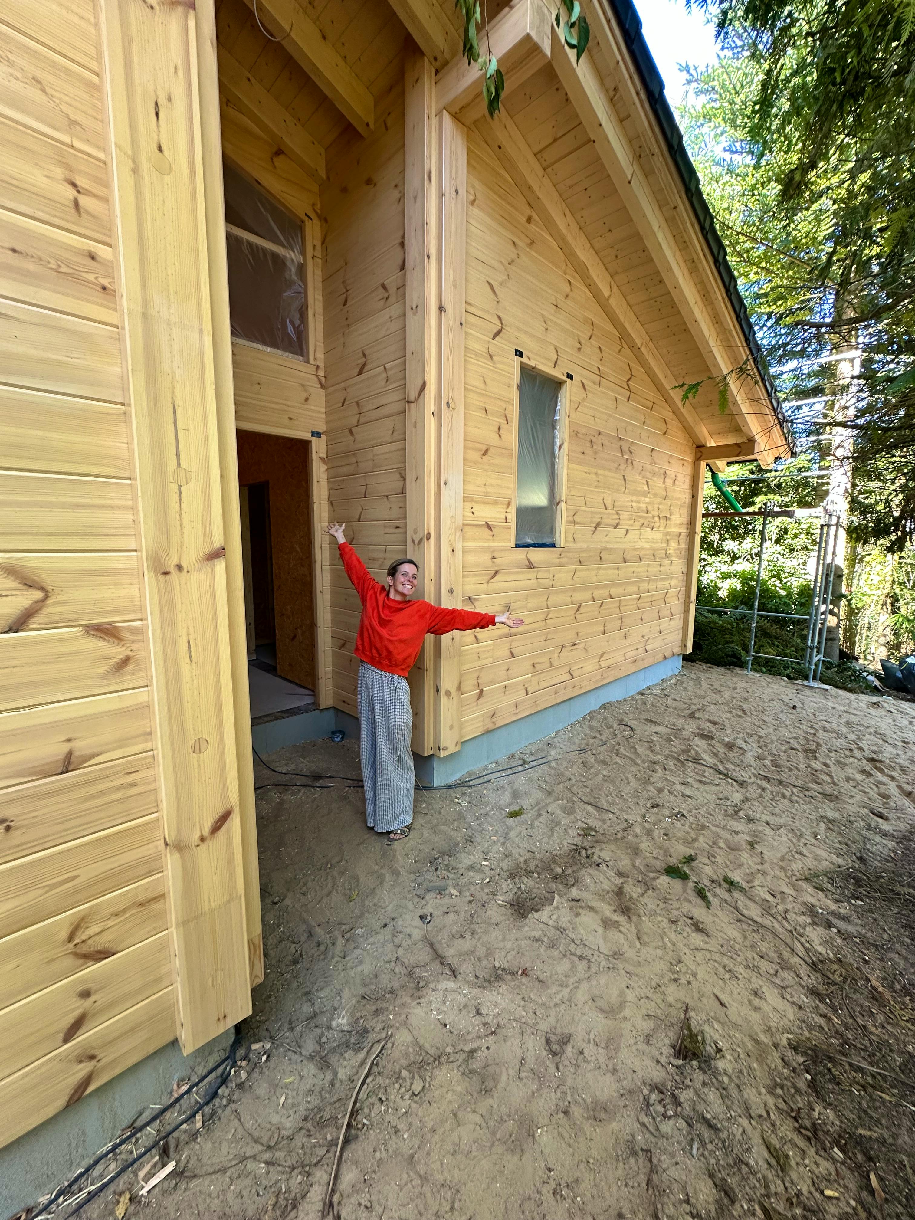 Bauherrin Claudia steht mit ausgebreiteten Armen vor der Polarkiefer-Fassade ihres Fullwood-Blockhauses