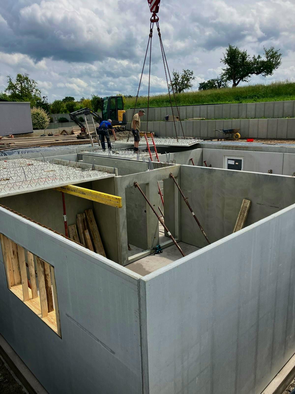 Fertigkeller im Aufbau: Betonwände stehen, ein Wandelement wird per Kran eingesetzt, zwei Arbeiter stehen auf der Kellerdecke, im Hintergrund grüne Felder