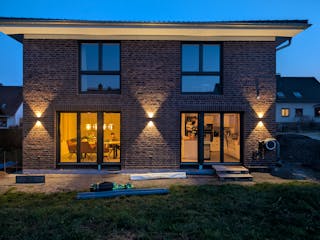 Rückansicht der Viebrockhaus-Stadtvilla in Bodenfelde bei Abenddämmerung – bodentiefe Fenster beleuchtet, Klinkerfassade mit Wandleuchten, Blick in den offenen Wohn-Essbereich