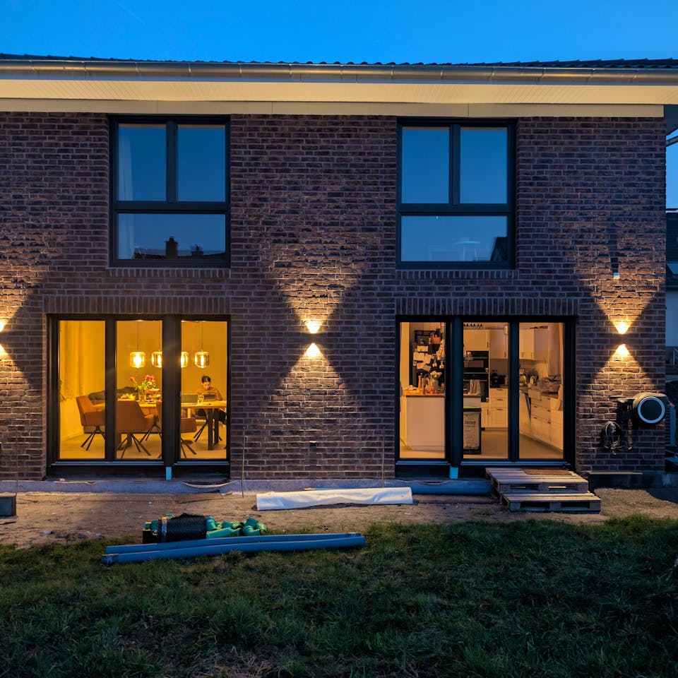Rückansicht der Viebrockhaus-Stadtvilla in Bodenfelde bei Abenddämmerung – bodentiefe Fenster beleuchtet, Klinkerfassade mit Wandleuchten, Blick in den offenen Wohn-Essbereich