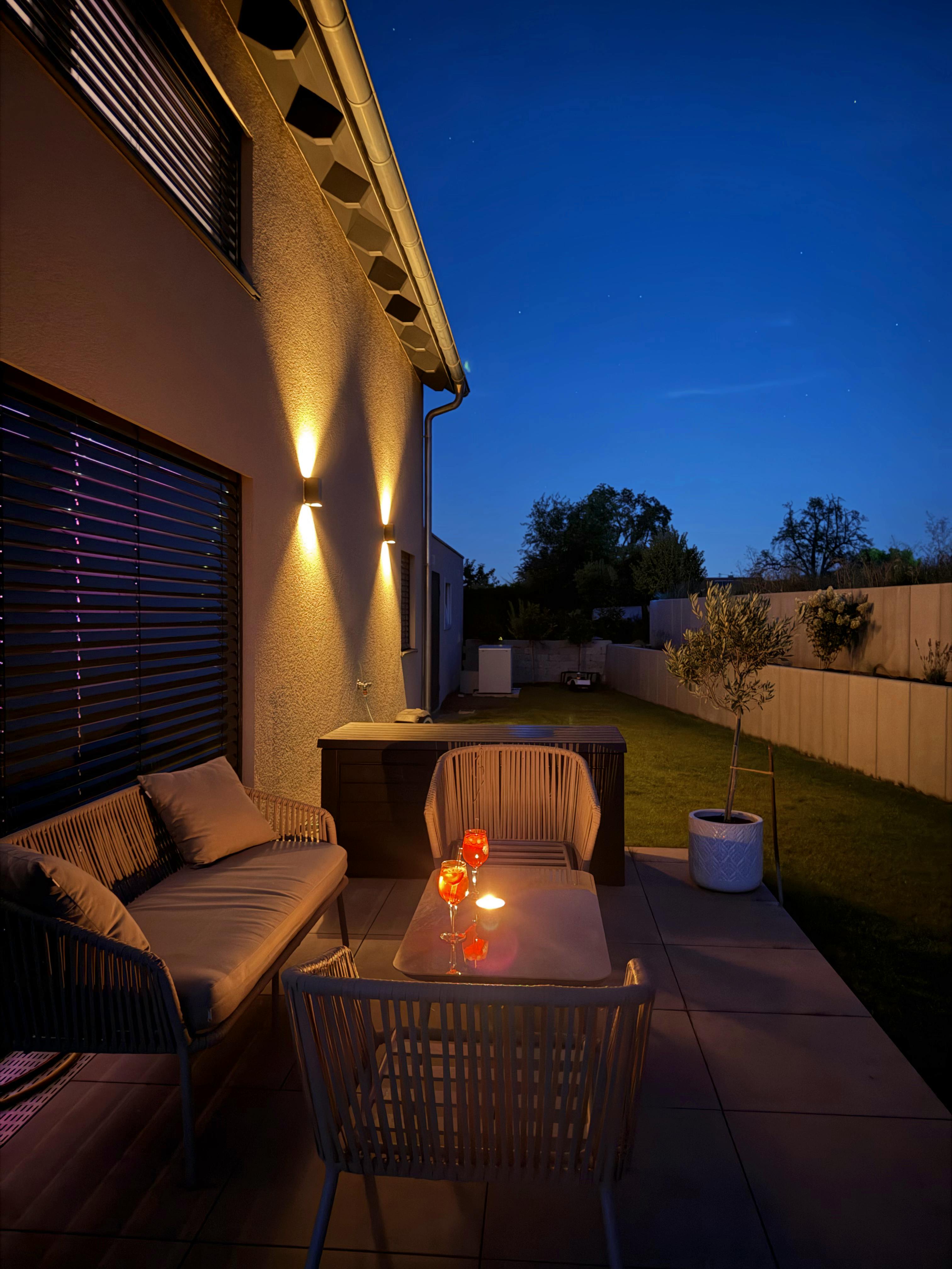 Terrasse bei Nacht mit warmweißen Wandleuchten, Lounge-Möbeln und zwei Cocktails