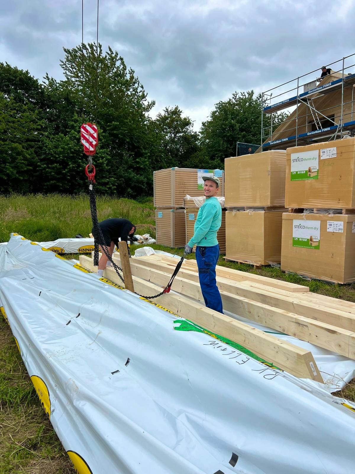 Helfer am Materiallager mit verpackten Finnholz-Blockbohlen und Steico-Dämmplatten, Baukran-Haken sichtbar