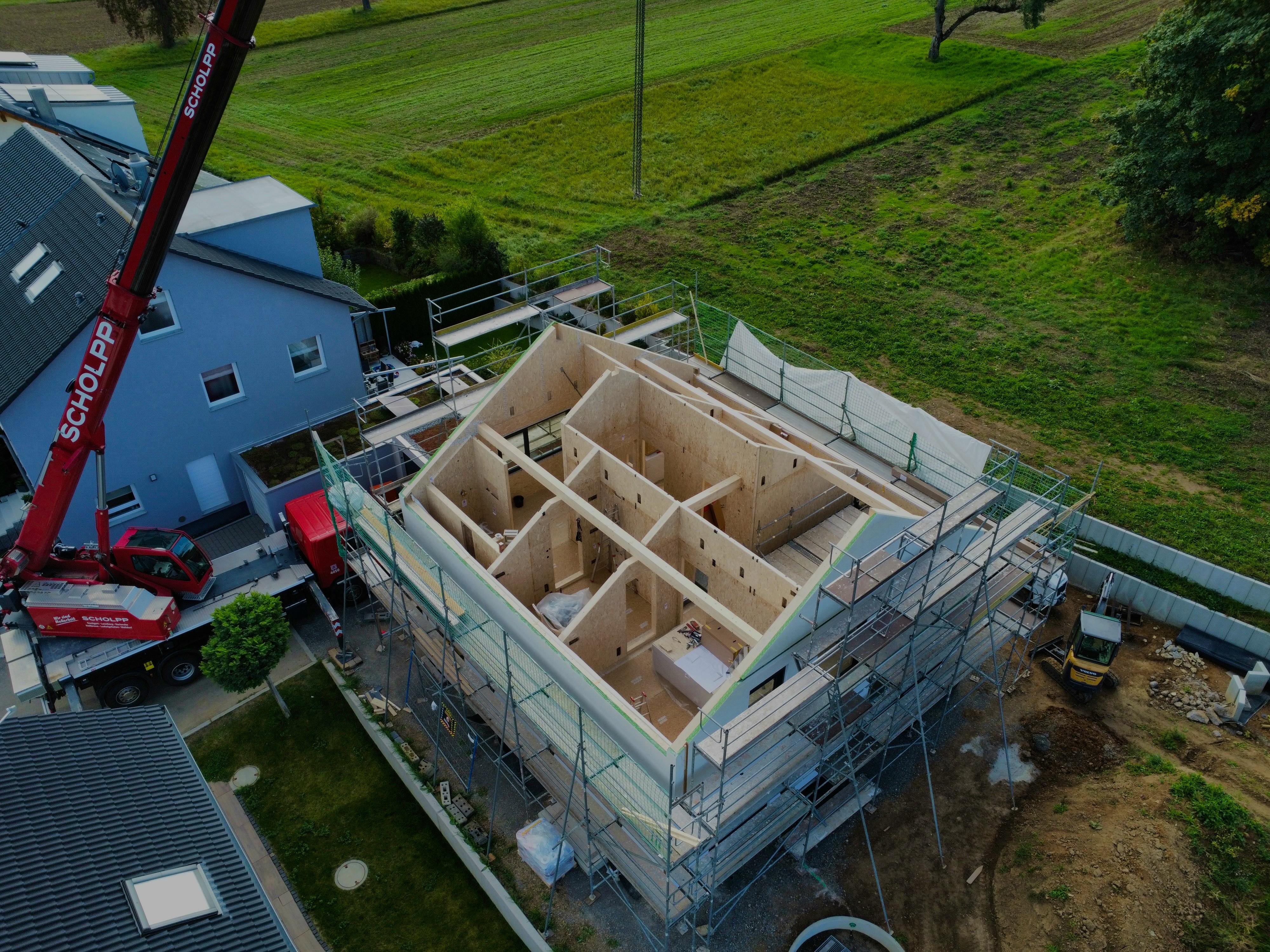 Drohnenaufnahme des LUXHAUS-Rohbaus während des Stelltags, Holzständerbauweise sichtbar, Kran und grüne Felder im Hintergrund