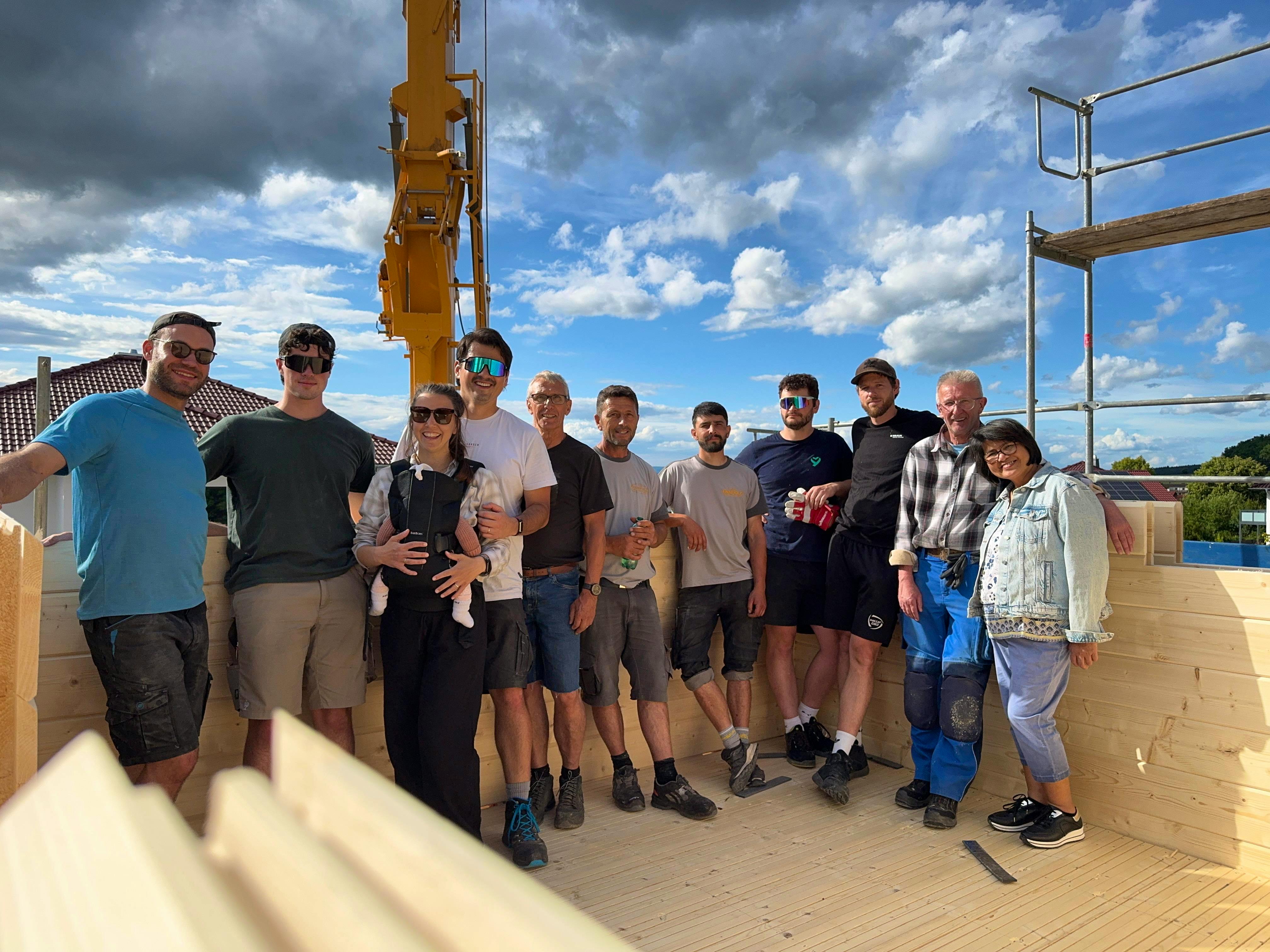 Gruppenfoto des Helfer-Teams mit Tim, Annemarie mit Baby, Richtmeistern und Freunden im Obergeschoss des Blockhauses, Baukran im Hintergrund
