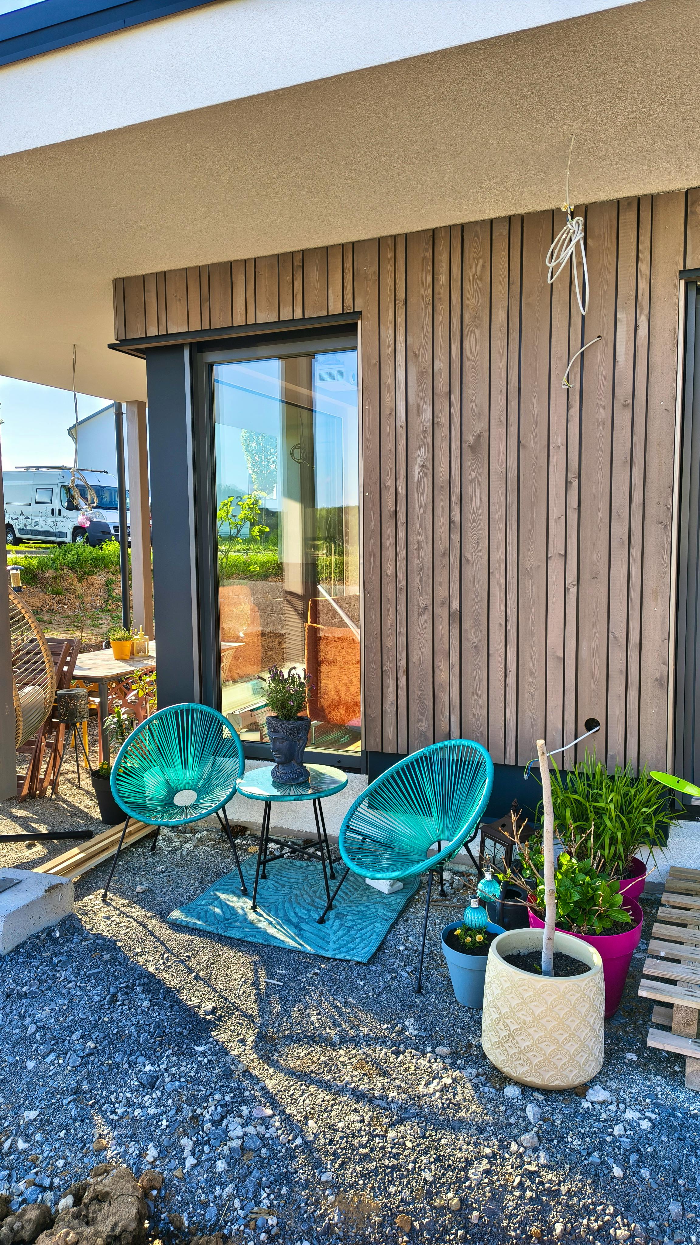 Nahaufnahme der Holzfassade unter der Terrassenüberdachung – vertikale Fichtenbretter, graue Fensterrahmen, provisorische Terrasse mit türkisen Stühlen