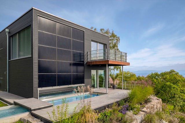 Designervilla bauen mit Bau-Fritz: Solarpanels an der Gebäudeseite sorgen für Energieeffizienz und einen optischen Hingucker, genau wie  die dunkle Holzverschalung in Kombination mit Stahl und einem Wasserbecken.